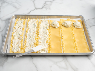 A baking sheet with six lasagna noodles laid flat. The left three are spread with ricotta cheese filling, while the right three have scoops of filling—perfect prep for a Lasagna Roll Ups recipe. A spatula rests on the tray.