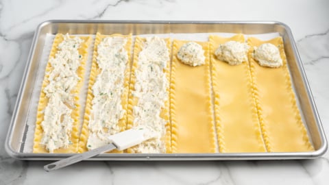 A baking sheet with six lasagna noodles laid flat. The left three are spread with ricotta cheese filling, while the right three have scoops of filling&mdash;perfect prep for a Lasagna Roll Ups recipe. A spatula rests on the tray.