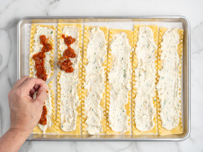 A hand spreads red tomato sauce onto a cooked lasagna noodle lined with ricotta cheese mixture, while several other noodles for the Lasagna Roll Ups are arranged on a baking sheet, ready to complete this delicious lasagna roll ups recipe.