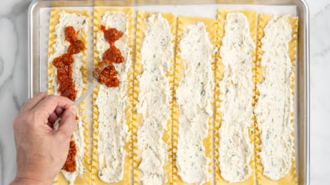 A hand spreads red tomato sauce onto a cooked lasagna noodle lined with ricotta cheese mixture, while several other noodles for the Lasagna Roll Ups are arranged on a baking sheet, ready to complete this delicious lasagna roll ups recipe.