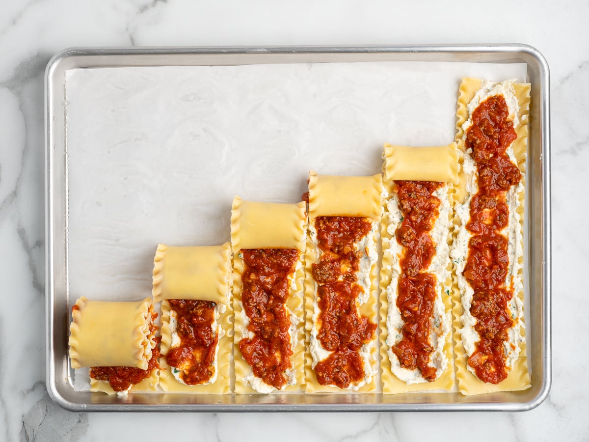 Six lasagna noodles are lined up on a baking sheet, each topped with ricotta cheese and tomato sauce, showing the step-by-step process of making this tasty Lasagna Roll Ups recipe.