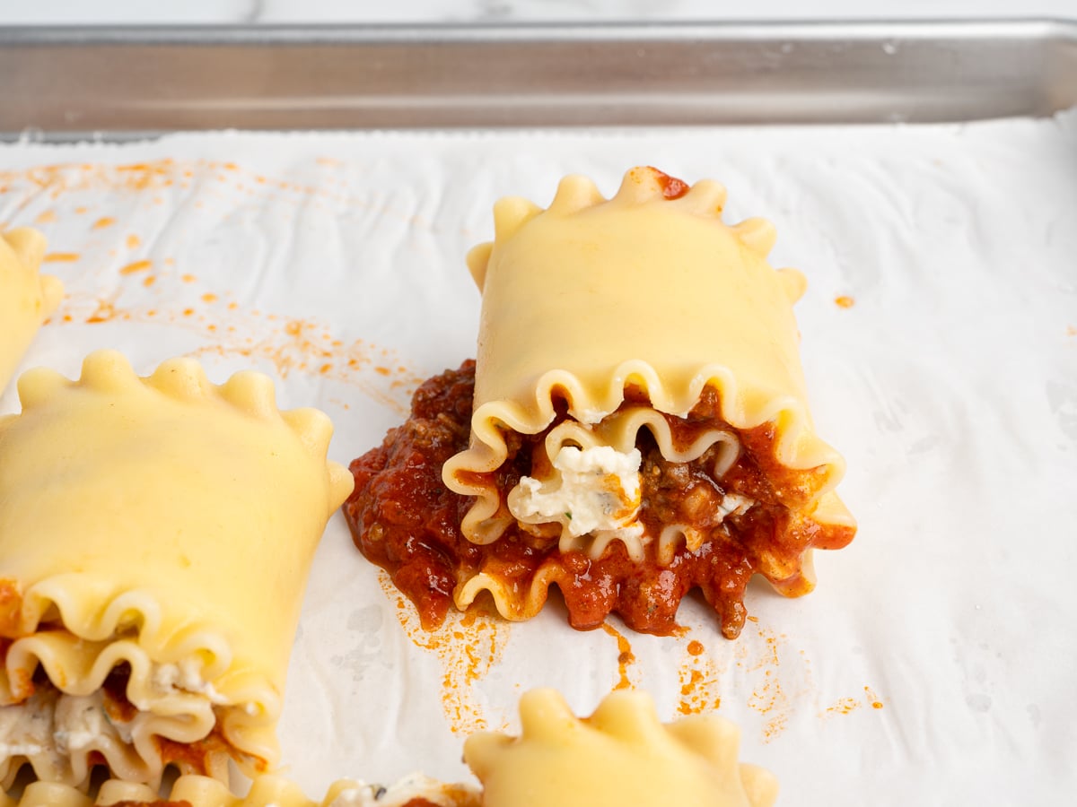 Three Lasagna Roll Ups sit on a parchment-lined baking sheet. One roll-up is spilling out with ricotta cheese and tomato meat sauce, while the others remain neatly rolled—an irresistible lasagna roll ups recipe in the making.