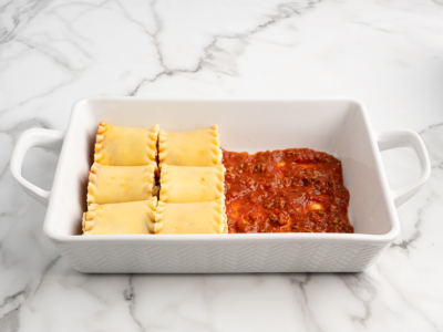 A white baking dish on a marble surface contains four uncooked Lasagna Roll Ups on one side and a layer of red tomato sauce with ground meat on the other, ready for the next step in this delicious lasagna roll ups recipe.
