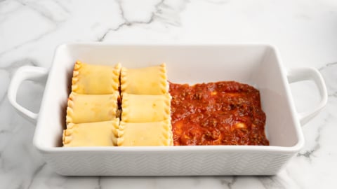 A white baking dish on a marble surface contains four uncooked Lasagna Roll Ups on one side and a layer of red tomato sauce with ground meat on the other, ready for the next step in this delicious lasagna roll ups recipe.