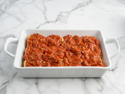 A white rectangular baking dish contains four Lasagna Roll Ups topped with chunky red meat sauce, sitting on a white marble surface.