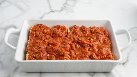 A white rectangular baking dish contains four Lasagna Roll Ups topped with chunky red meat sauce, sitting on a white marble surface.
