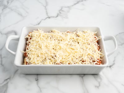 A white rectangular baking dish with handles, filled with Lasagna Roll Ups topped with a generous layer of shredded cheese, sits on a white marble countertop.