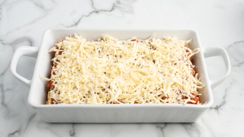 A white rectangular baking dish with handles, filled with Lasagna Roll Ups topped with a generous layer of shredded cheese, sits on a white marble countertop.