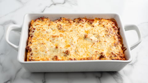A rectangular white baking dish filled with delicious Lasagna Roll Ups, topped with melted golden-brown cheese, sits on a white marble surface.