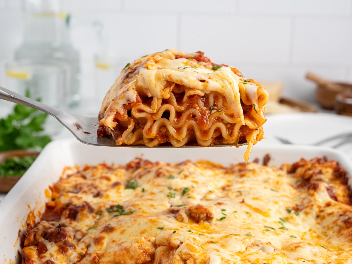 A close-up of a cheesy slice of lasagna being lifted from a baking dish, showing layers of pasta, meat sauce, and melted cheese—perfect inspiration for your next Lasagna Roll Ups recipe. More lasagna sits temptingly in the dish below.
