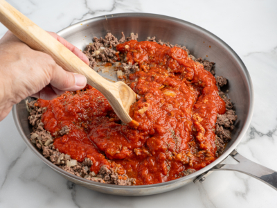 A hand stirs tomato sauce into cooked ground beef in a stainless steel skillet with a wooden spoon on a marble countertop, preparing the savory filling for a delicious Lasagna Roll Ups recipe.