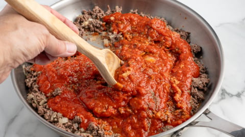 A hand stirs tomato sauce into cooked ground beef in a stainless steel skillet with a wooden spoon on a marble countertop, preparing the savory filling for a delicious Lasagna Roll Ups recipe.