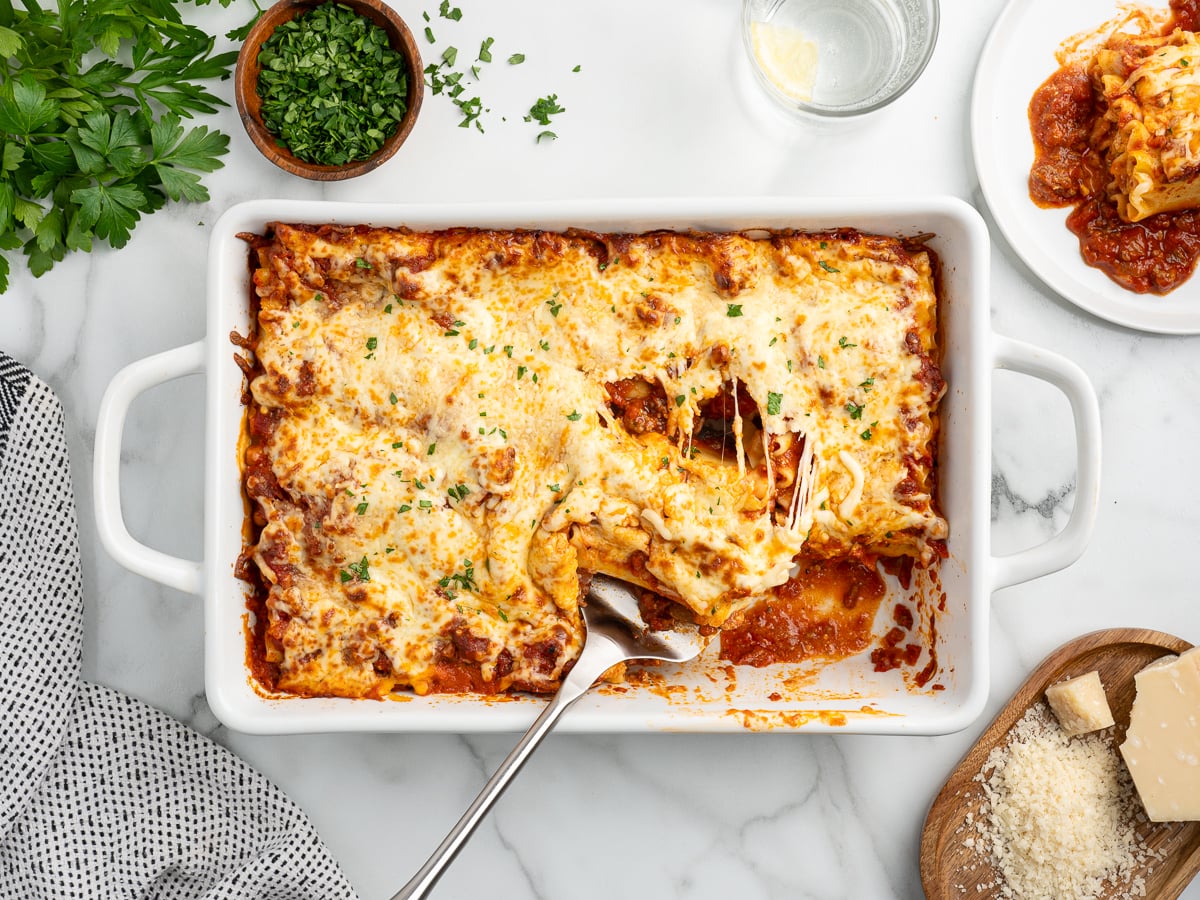 A baked Lasagna Roll Ups dish in a white casserole, with a serving spoon lifting one cheesy piece. The lasagna is topped with melted cheese and herbs, surrounded by fresh parsley, grated cheese, a glass of water, and a serving plate.