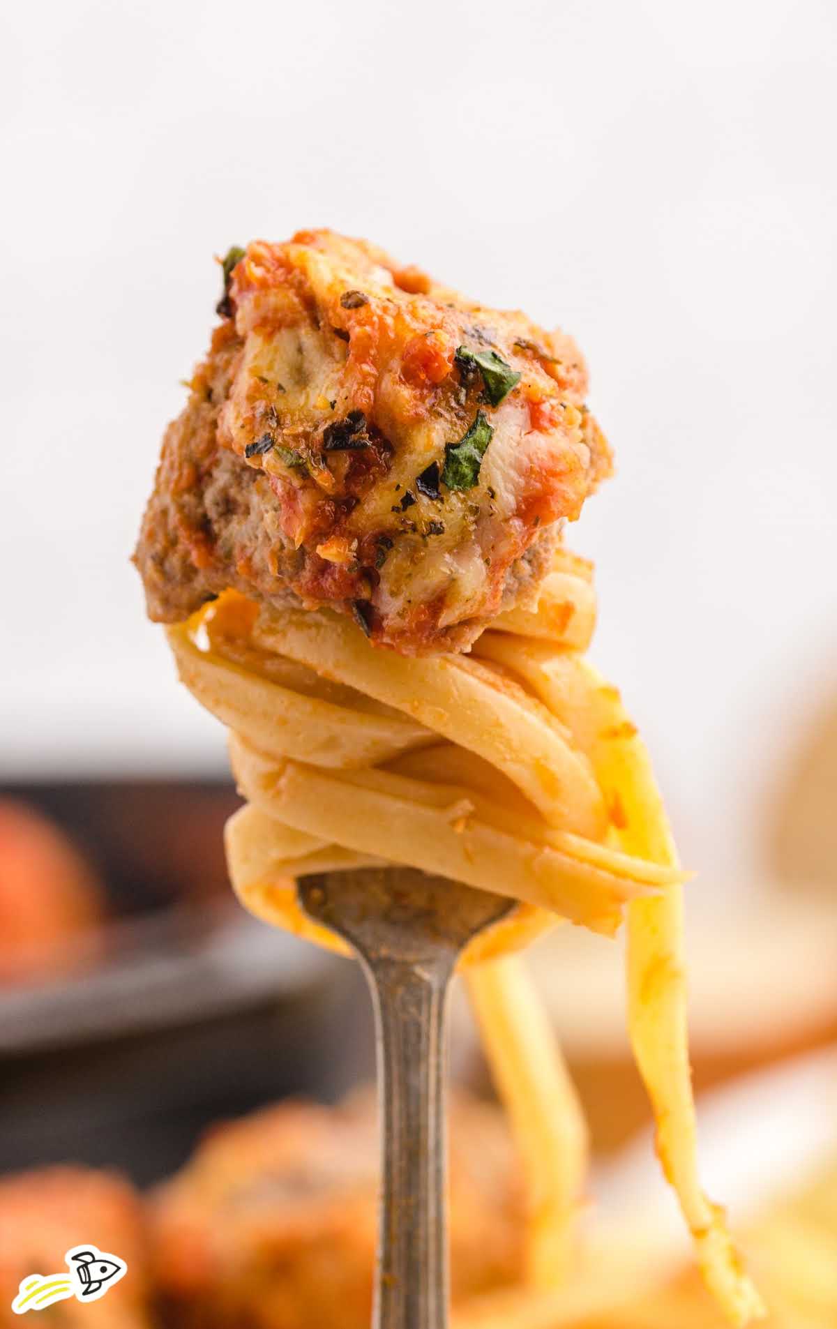 Fork twirling pasta topped with a single baked meatball in front of a blurred skillet background.