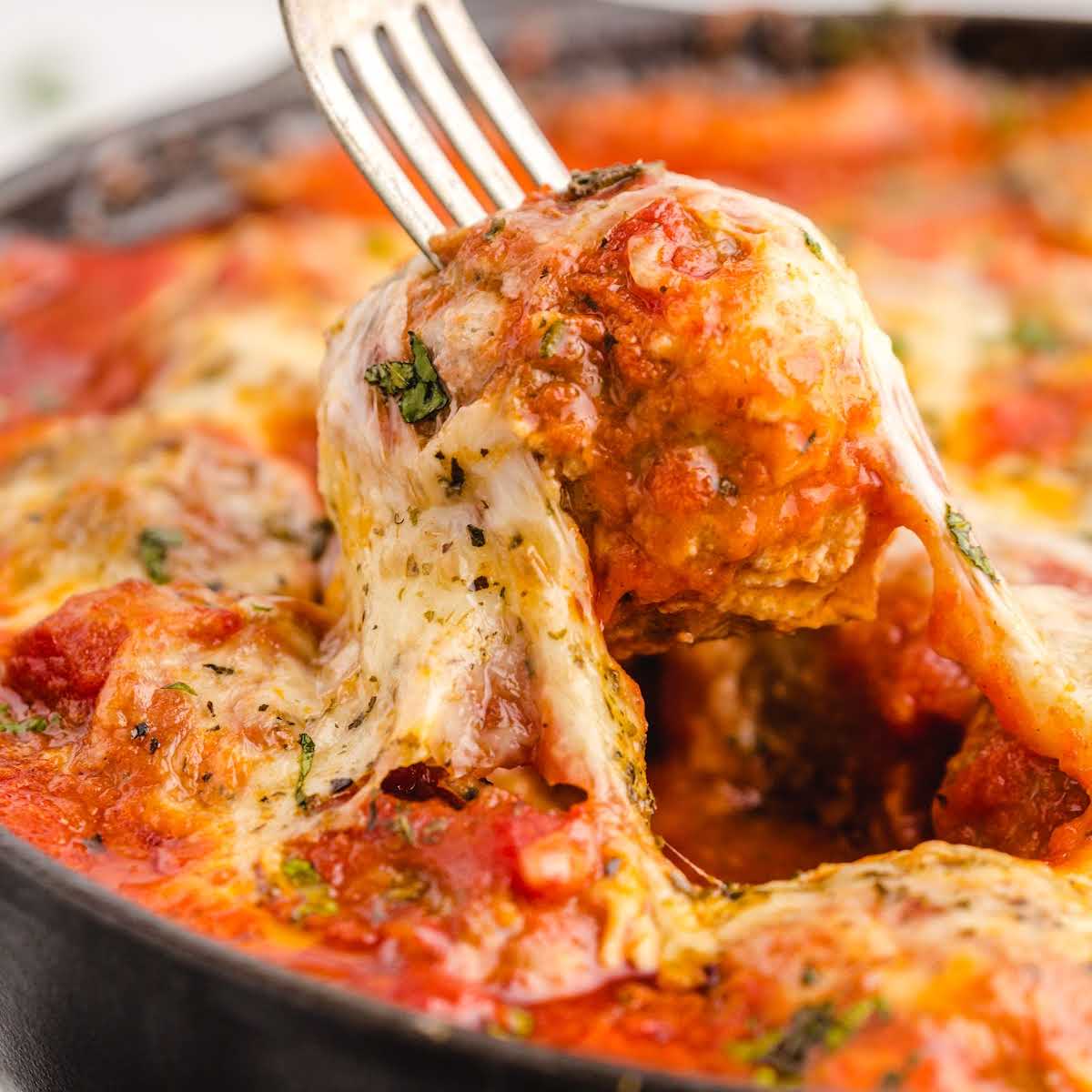 Square close-up of a cheesy meatball being pulled from a skillet with a fork, showcasing melted mozzarella and vibrant marinara sauce.