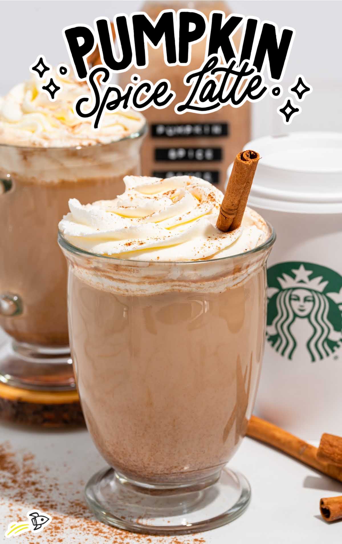Pumpkin spice latte in a glass mug topped with whipped cream and a cinnamon stick, with pumpkin spice sauce bottle in background.