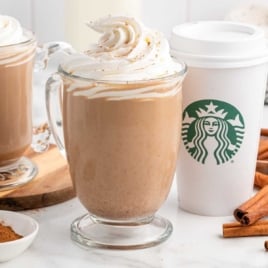 Close-up of a glass mug filled with pumpkin spice latte and topped with whipped cream, next to a Starbucks to-go cup.
