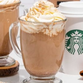 Two pumpkin spice lattes with whipped cream in clear glass mugs, surrounded by cinnamon sticks and Starbucks cups.