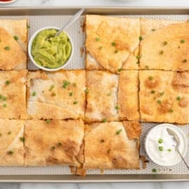 close up overhead shot of quesadillas with cups of salsa, sour cream, and guacamole on a sheet pan