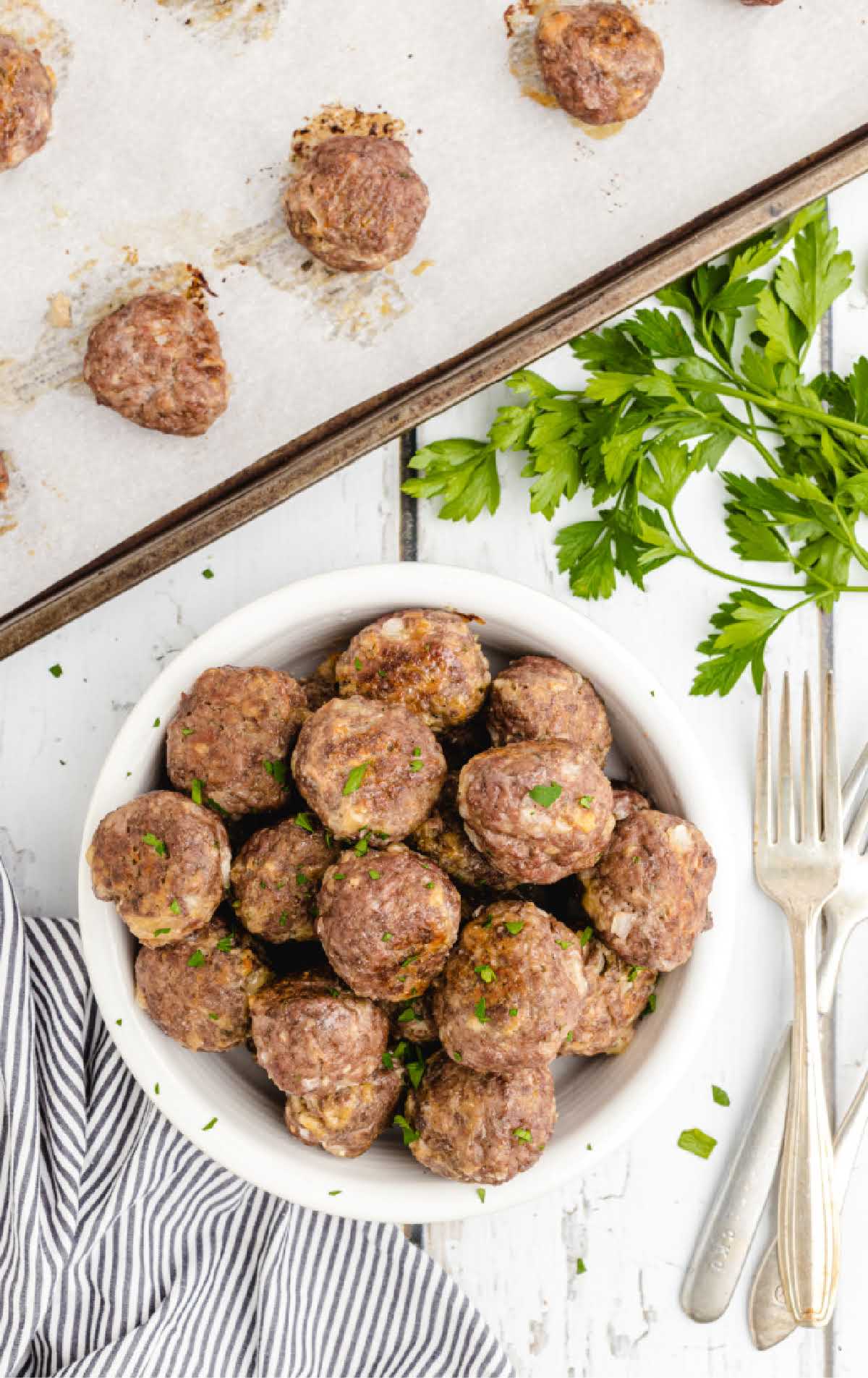 Baked meatballs on a parchment-lined baking sheet and a bowl of cooked meatballs on a white surface.