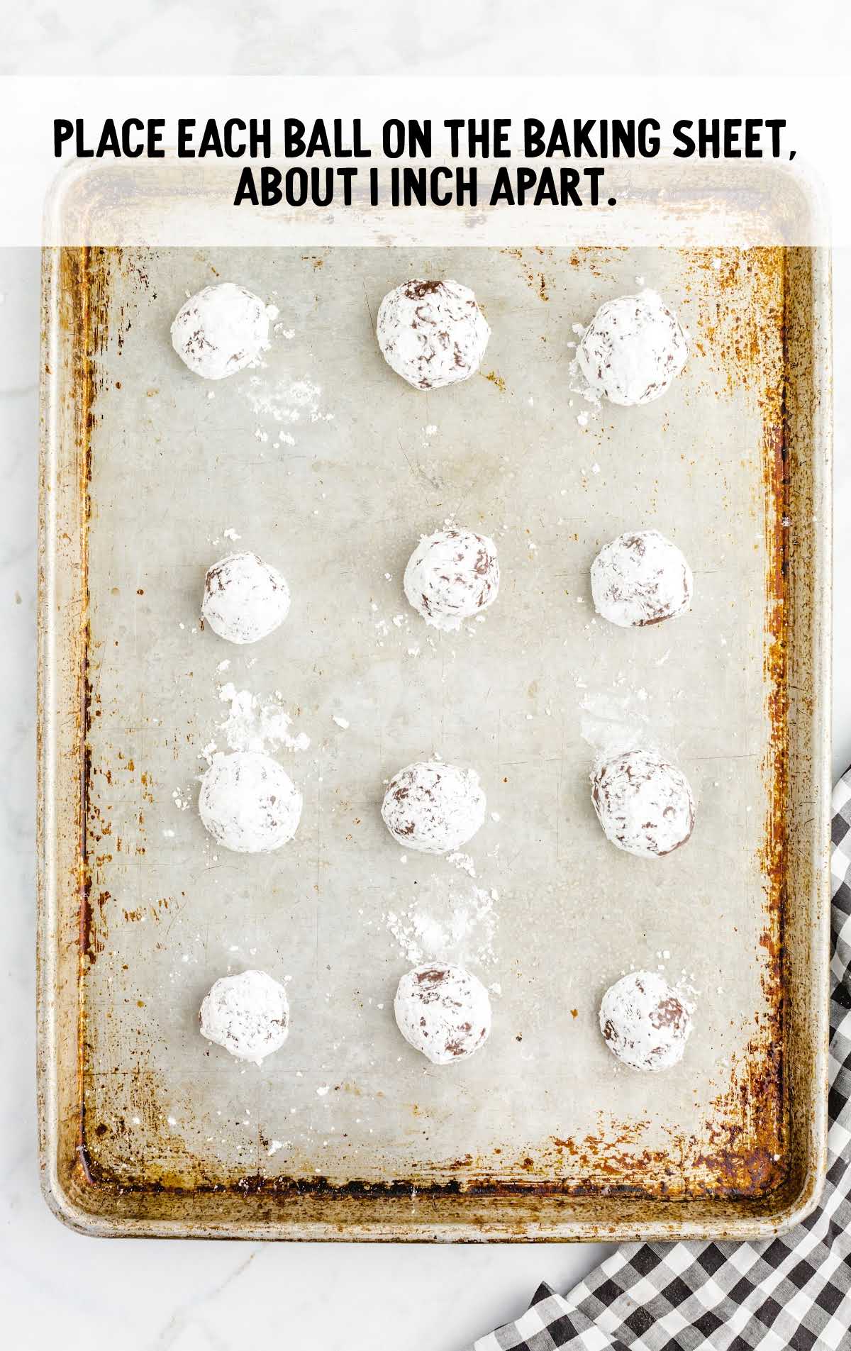 Powdered sugar-coated cookie dough balls spaced evenly on a baking sheet, with text overlay: “Place each ball on the baking sheet, about 1 inch apart.”
