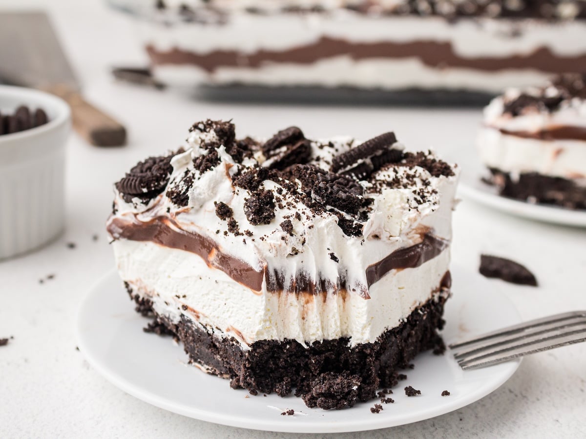 A slice of Oreo Delight dessert with a chocolate cookie crust, creamy white filling, chocolate pudding layer, and whipped topping, garnished with crushed Oreos, sits on a white plate with a fork.