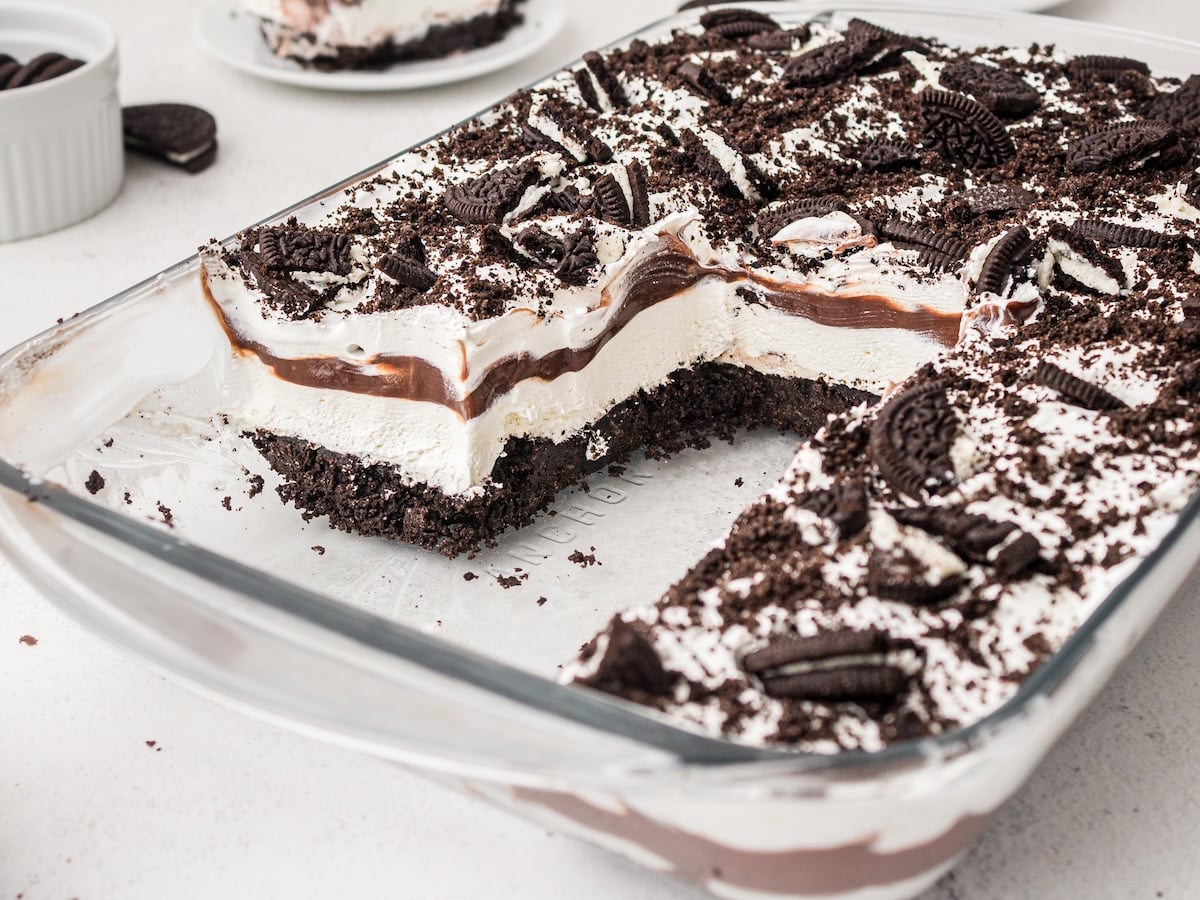 A glass dish of Oreo Delight Dessert with a chocolate cookie crumb base, creamy white filling, chocolate pudding layer, and whipped topping, finished with crushed and halved Oreos. A large slice of this Oreo delight with chocolate pudding has been removed.