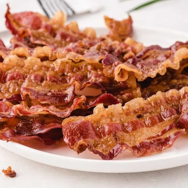 Side view of cooked, wavy bacon strips piled high on a white plate with a napkin and fork in the background.