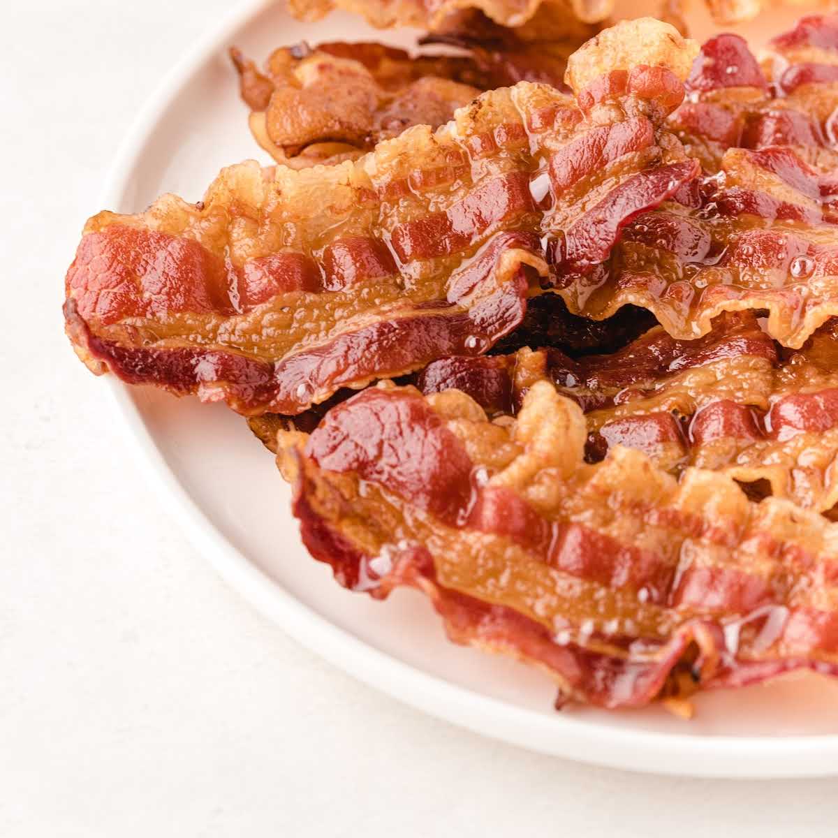 Text overlay "How to Cook Bacon in the Oven" over a close-up of glossy, golden strips of bacon on a plate.