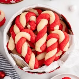 Round tin lined with white parchment paper filled with red and white twisted treats made using a classic Candy Cane Cookies Recipe, with more candy cane cookies and festive decorations visible around the tin.