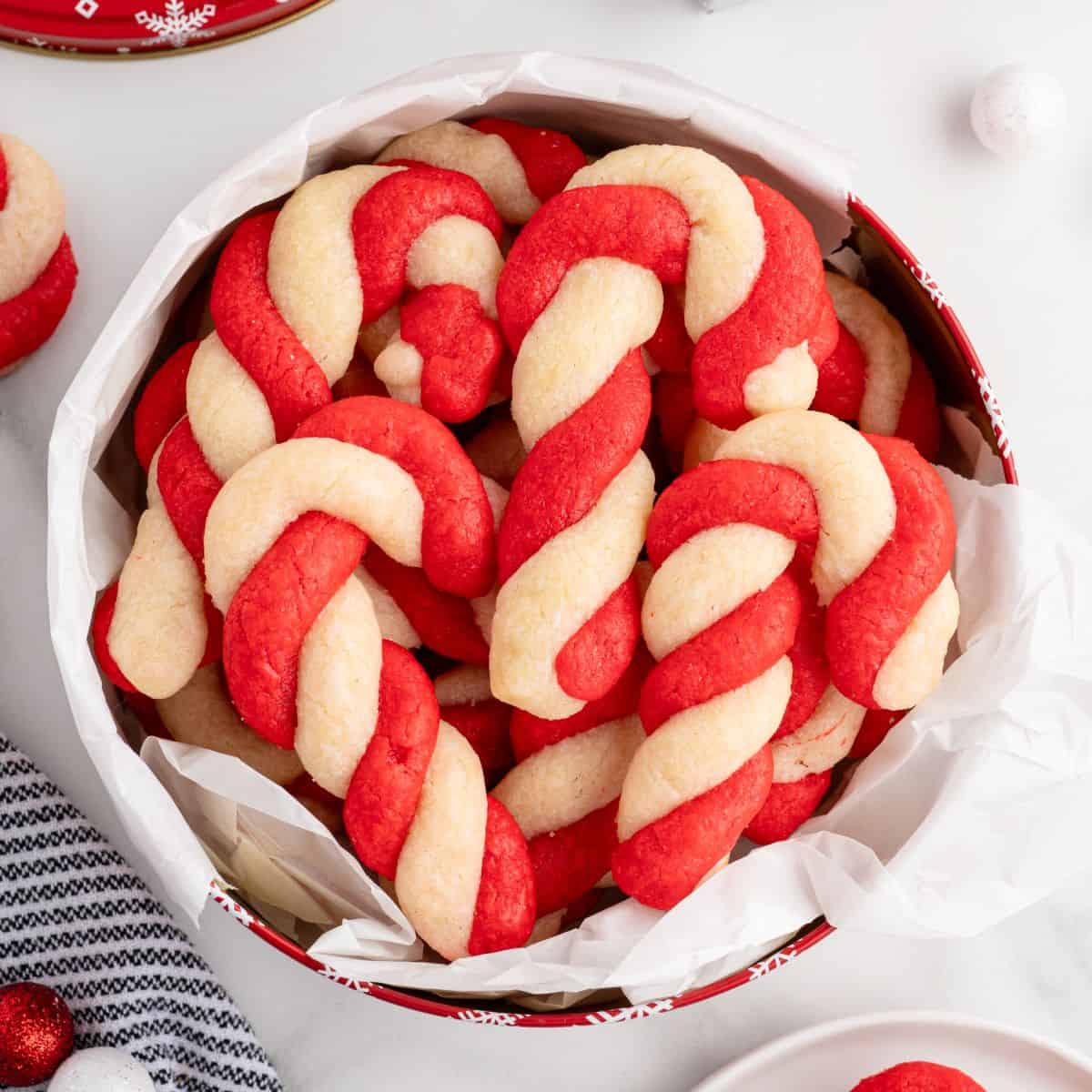 Round tin lined with white parchment paper filled with red and white twisted treats made using a classic Candy Cane Cookies Recipe, with more candy cane cookies and festive decorations visible around the tin.