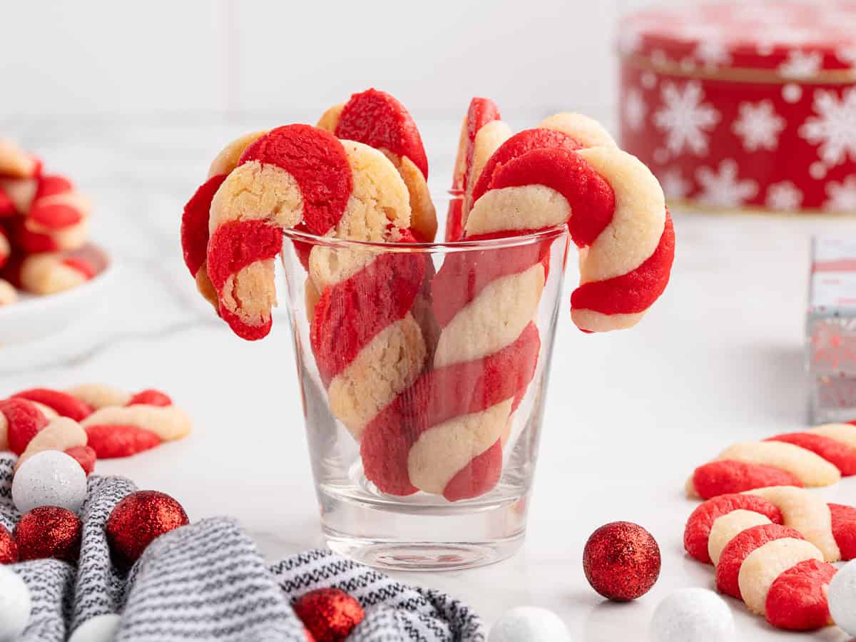 A clear glass holds festive Candy Cane Cookies, their red and white stripes curling just like classic holiday treats. More cookies and ornaments are scattered around on a white surface, with a holiday tin and plate completing this inviting scene—perfect inspiration for trying your own Candy Cane Cookies Recipe.