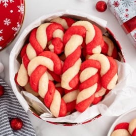 A round tin filled with red and white twisted Candy Cane Cookies rests on parchment paper, surrounded by festive holiday decorations and a wrapped gift—perfect for sharing or gifting after trying your favorite Candy Cane Cookies Recipe.