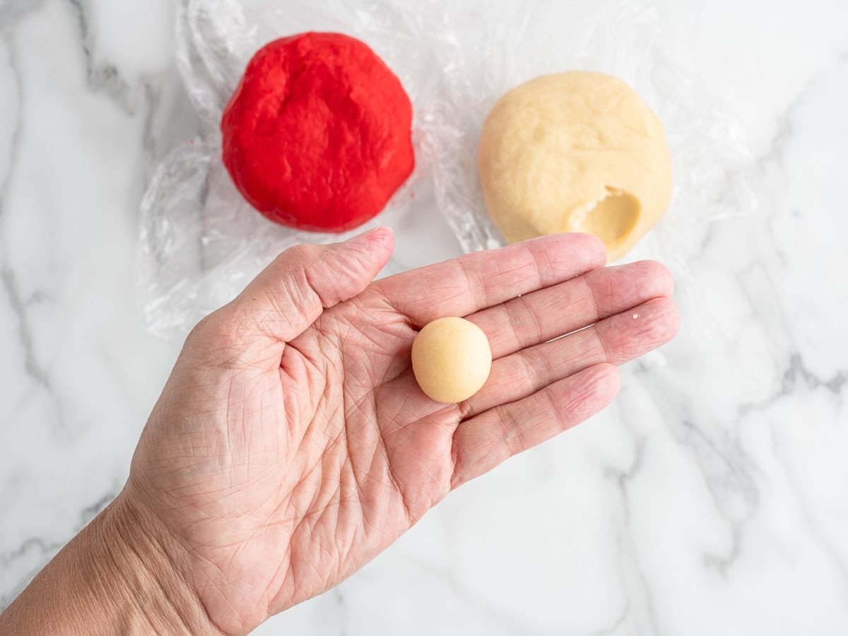 A hand holds a small ball of beige dough, perfect for shaping into Candy Cane Cookies. In the background, there are two larger balls of dough—one red and one beige—resting on plastic wrap on a marble surface, ready for the next step in your Candy Cane Cookies recipe.