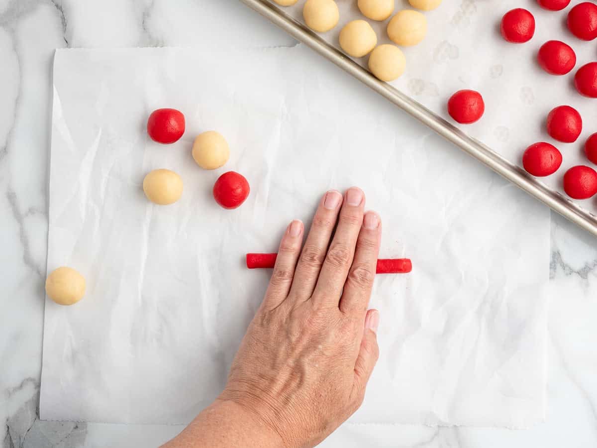 A hand rolls red dough into a log shape on parchment paper, beginning the process of making festive Candy Cane Cookies. Red and beige dough balls are nearby, with more shaped cookies on a baking sheet in the upper right corner. Marble countertop background.