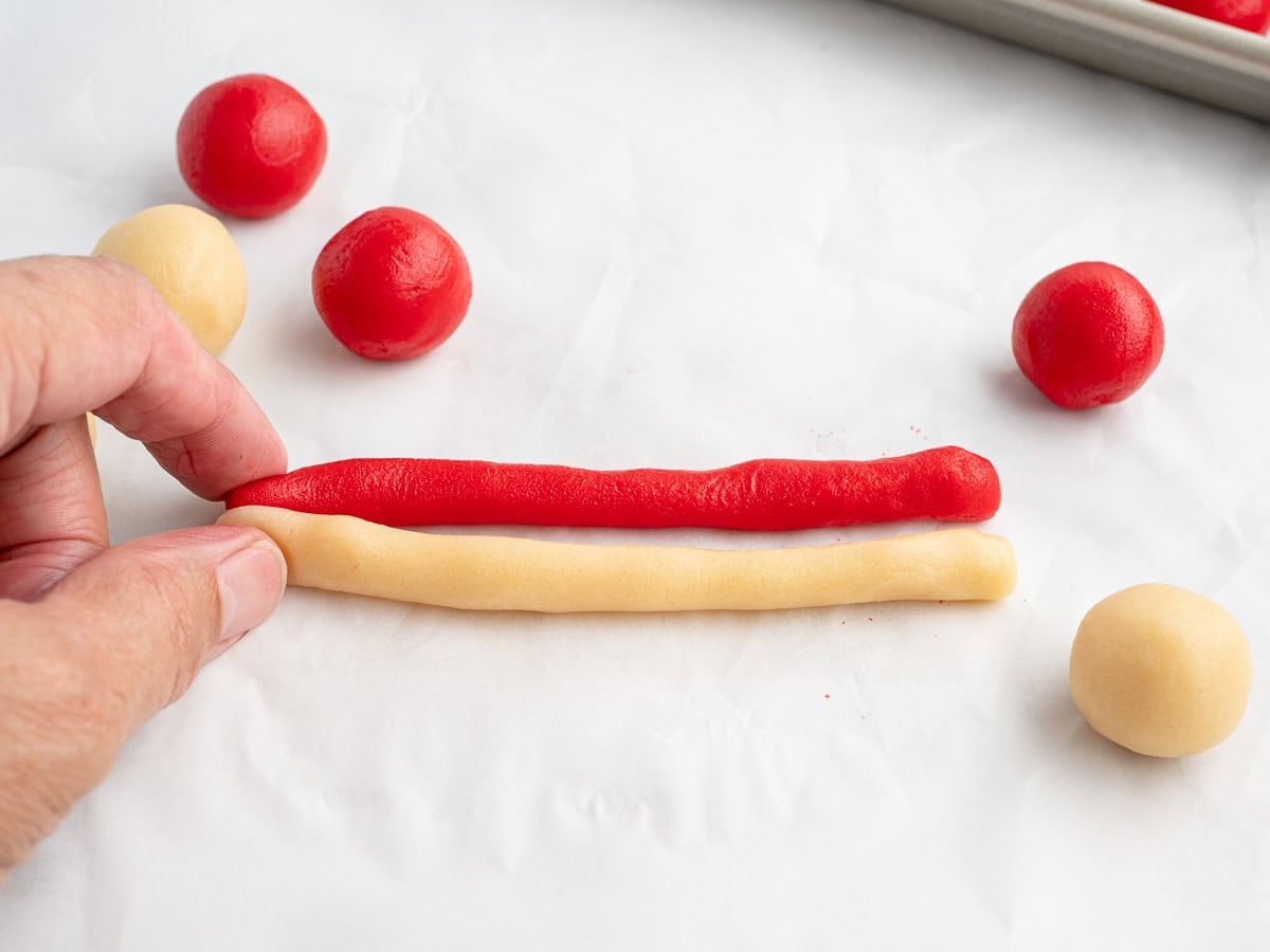 A hand shapes red and beige dough into long ropes on parchment paper, preparing to twist them for a festive Candy Cane Cookies Recipe, with round dough balls nearby and a baking sheet visible in the background.