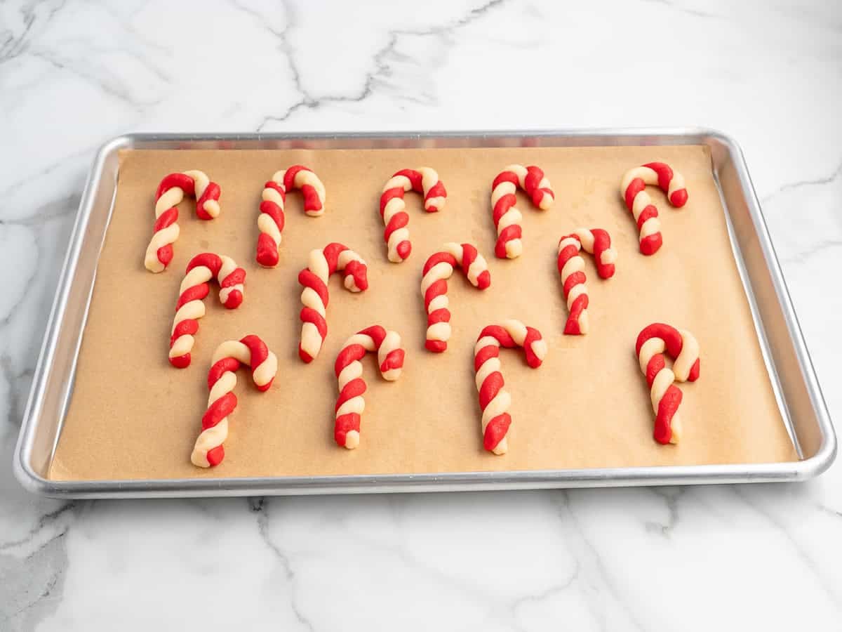 A baking sheet with parchment paper holds 16 delightful Candy Cane Cookies, twisted with red and white dough, on a marble countertop—perfect for anyone following a classic Candy Cane Cookies Recipe.