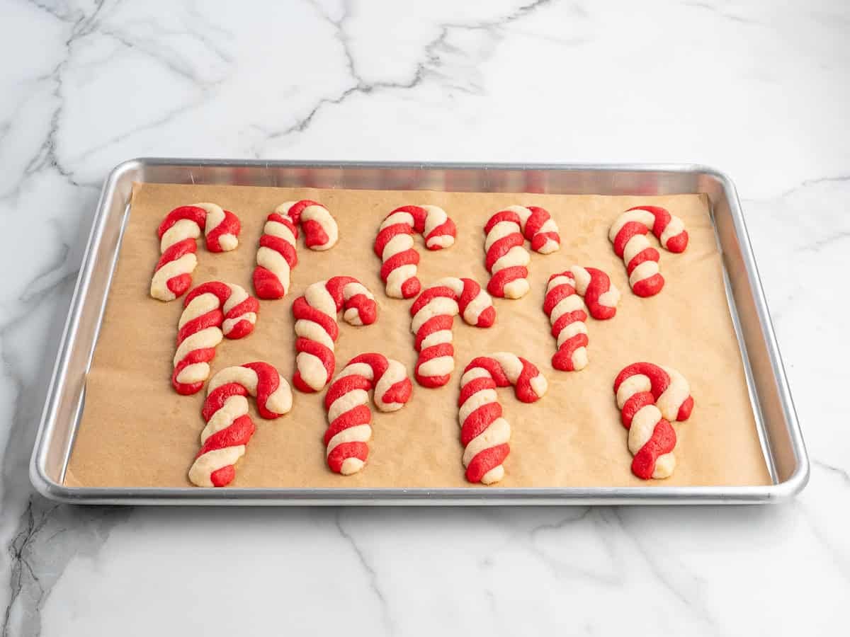 A baking tray lined with parchment paper holds several candy cane-shaped cookies with red and white twisted dough, resting on a marble countertop—an inviting scene inspired by the classic Candy Cane Cookies Recipe.