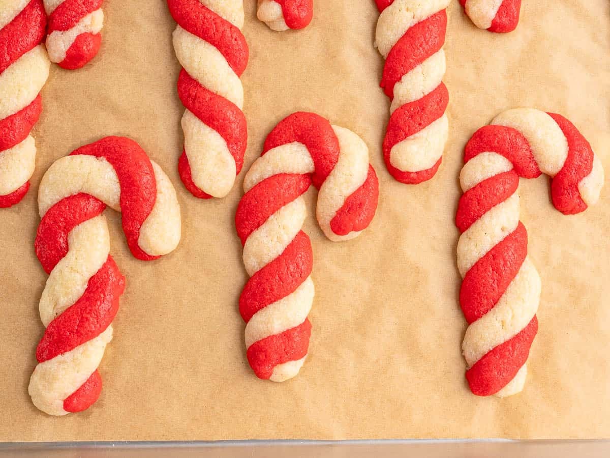 Candy Cane Cookies with red and white twisted dough are arranged on a sheet of brown parchment paper, ready to be baked or served.