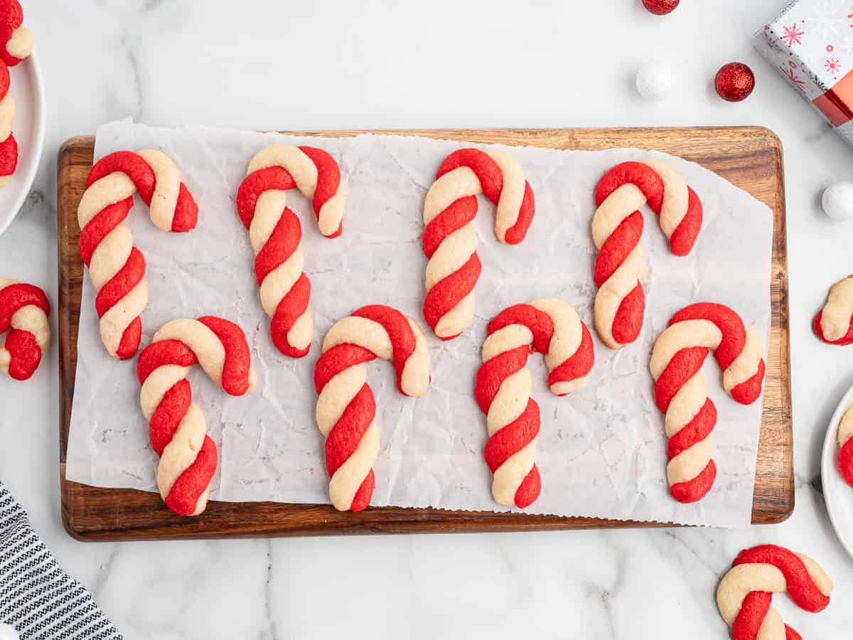 Nine red and white Candy Cane Cookies are arranged on parchment paper atop a wooden board, with holiday decorations and treats surrounding them on a marble surface.