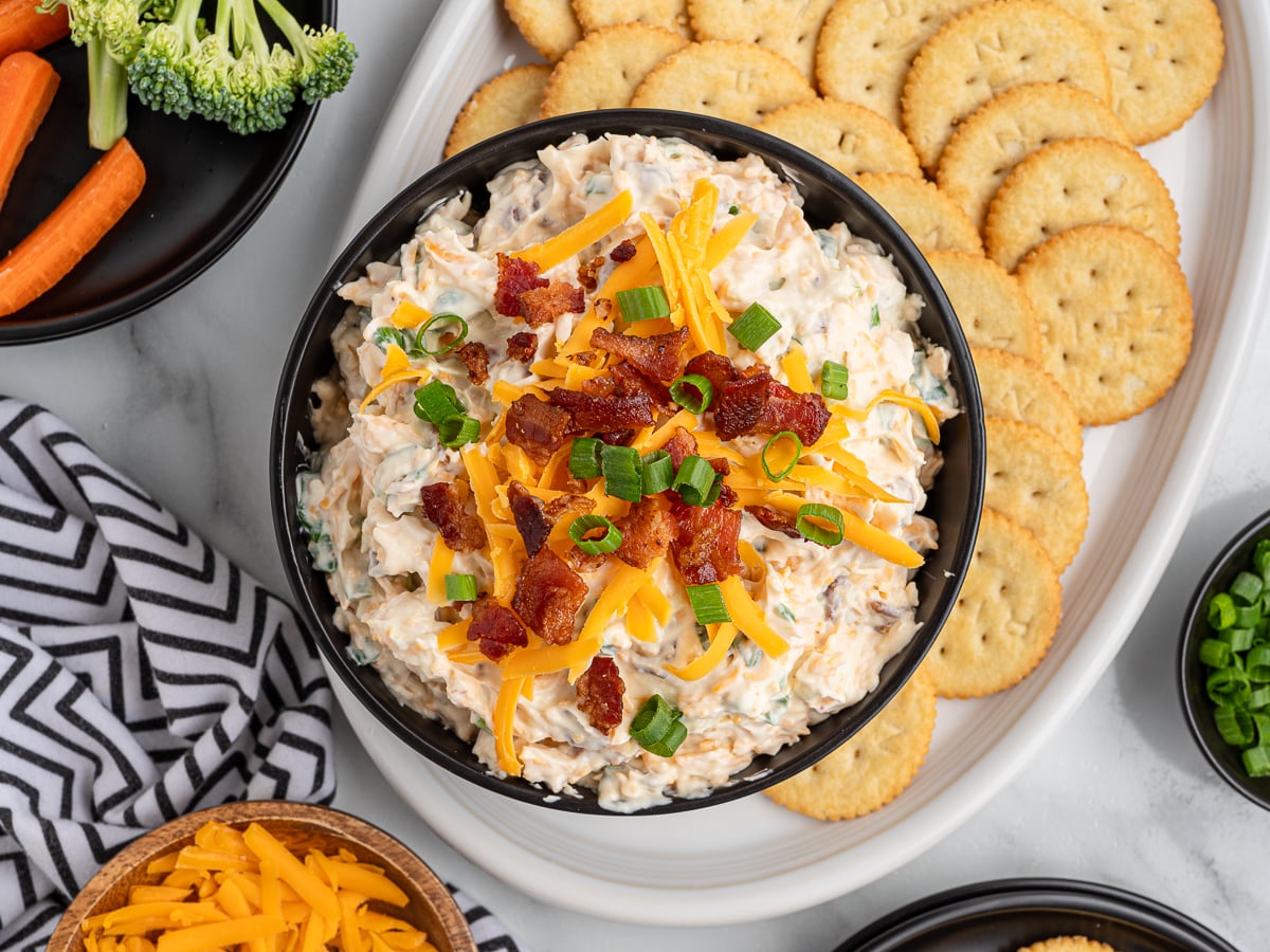 A bowl of creamy Million Dollar Dip topped with shredded cheddar cheese, bacon bits, and green onions, surrounded by round crackers, broccoli, carrots, and extra cheese on a white platter.