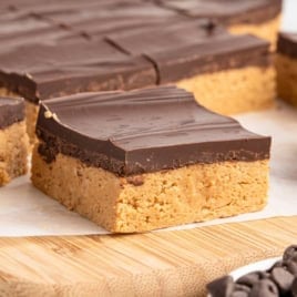 A close-up of no bake peanut butter bars featuring a thick peanut butter base and a smooth chocolate layer on top, sitting on a wooden board.