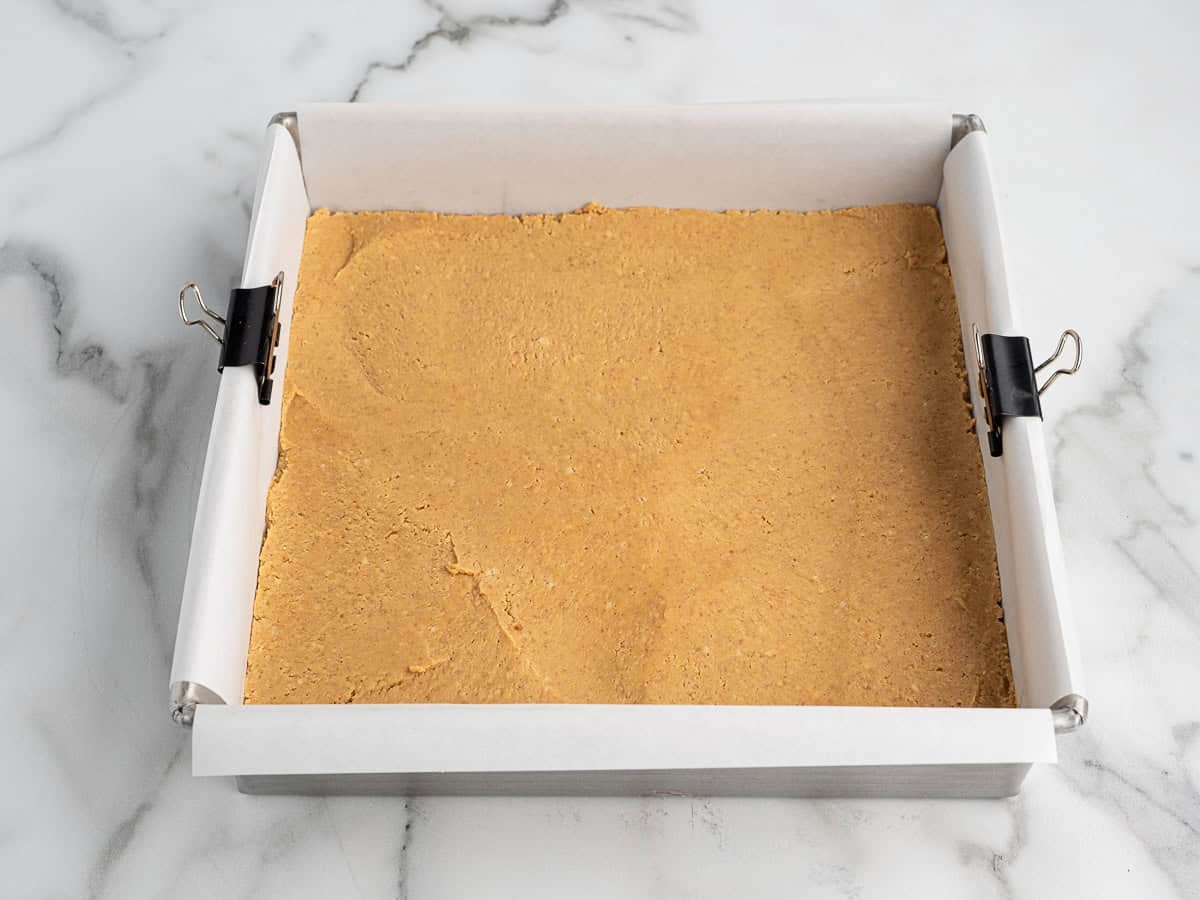 A square baking pan lined with parchment paper and binder clips, filled with a pressed layer of crumbly, light brown dough—perfect for making peanut butter squares—sits on a marble countertop.