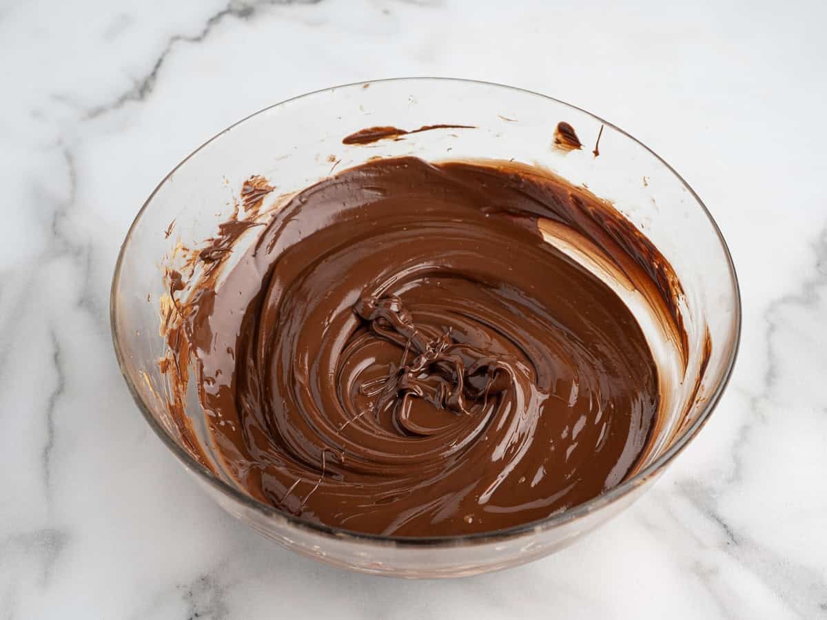 A clear glass bowl filled with smooth, melted chocolate sits on a white marble surface. Some chocolate is swirled around the sides of the bowl, ready to be poured over homemade peanut butter bars for an irresistible treat.