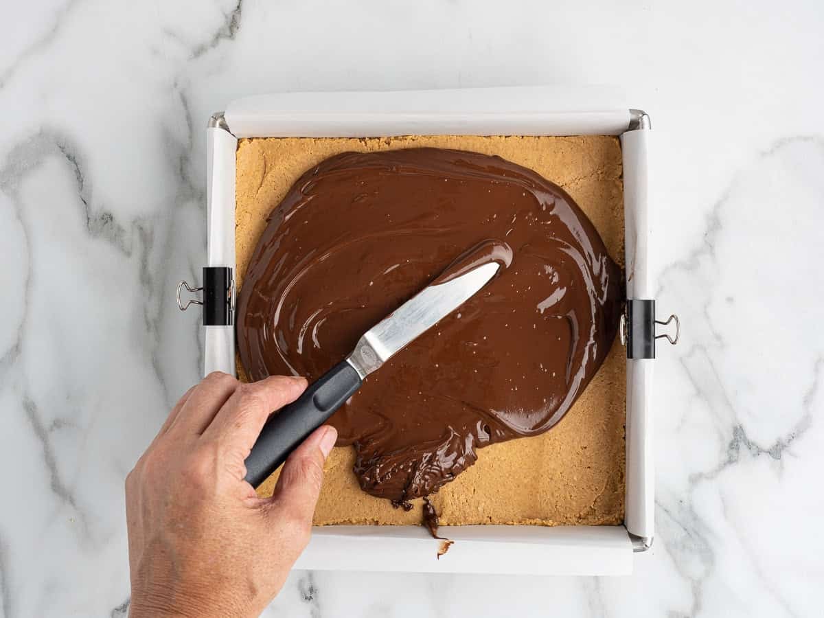 A hand spreads melted chocolate over a square pan of cookie crust lined with parchment paper, using a spatula on a marble countertop—an essential step in making irresistible chocolate peanut butter bars.
