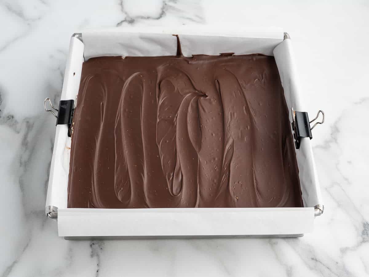 A square baking pan lined with parchment paper, filled with a smooth layer of melted chocolate for a classic peanut butter bars recipe, sits on a marble surface. Black binder clips hold the paper in place.