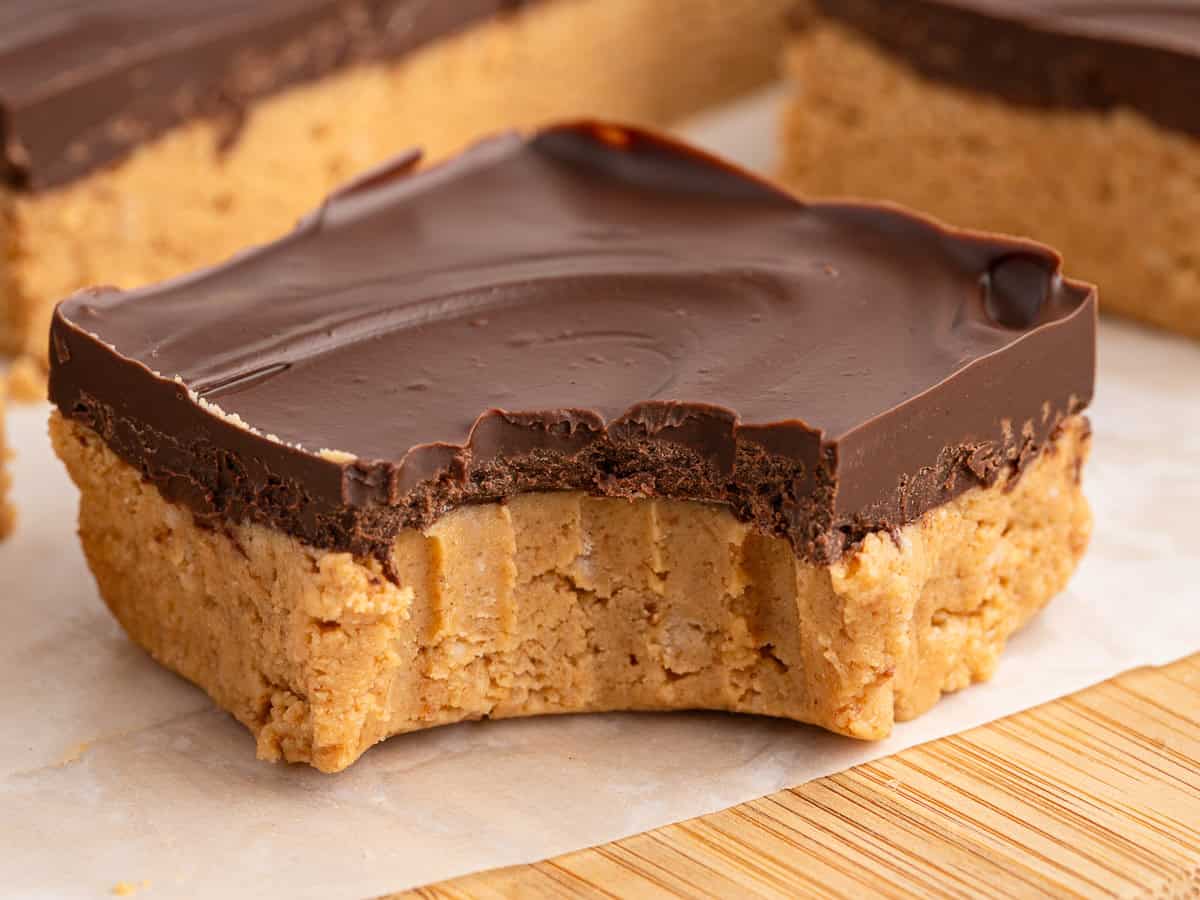 A close-up of a no bake peanut butter bar topped with chocolate, showing a large bite taken out of it. Other peanut butter squares are visible in the background on parchment paper.