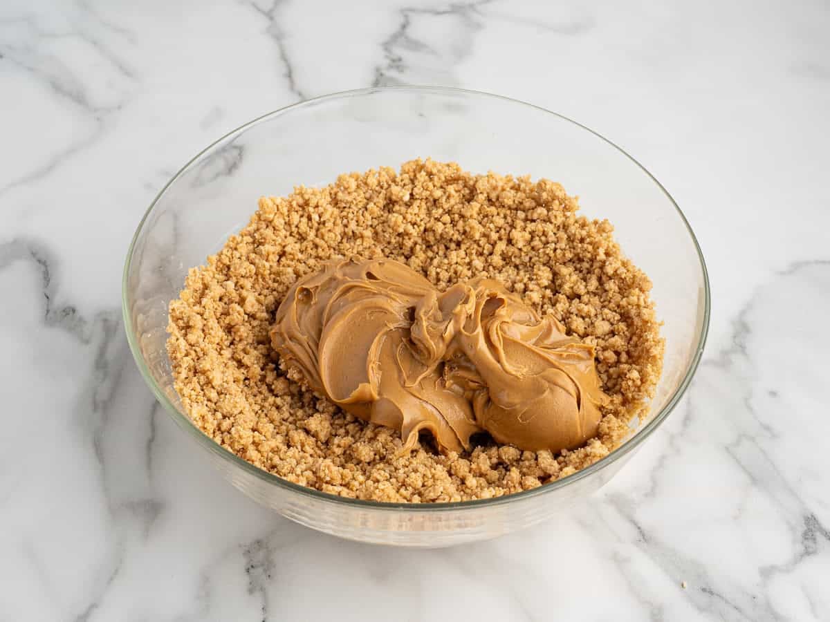 A glass bowl contains a mixture of crushed graham crackers and two large scoops of creamy peanut butter—perfect for making easy no bake peanut butter bars—on a white marble surface.