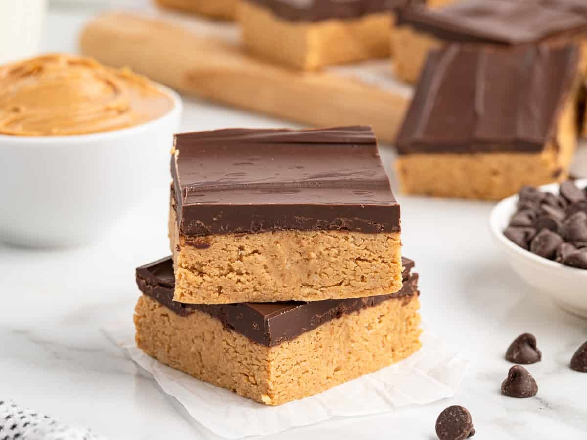 Two chocolate-topped peanut butter bars stacked on parchment paper, with more peanut butter bars, a bowl of creamy peanut butter, and scattered chocolate chips in the background—perfect for anyone craving an easy no bake peanut butter bars recipe.