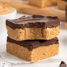 Two chocolate-topped peanut butter bars are stacked on parchment paper, with one showing a bite taken out—perfectly highlighting these irresistible no bake peanut butter bars. A bowl of peanut butter and more bars are in the background.